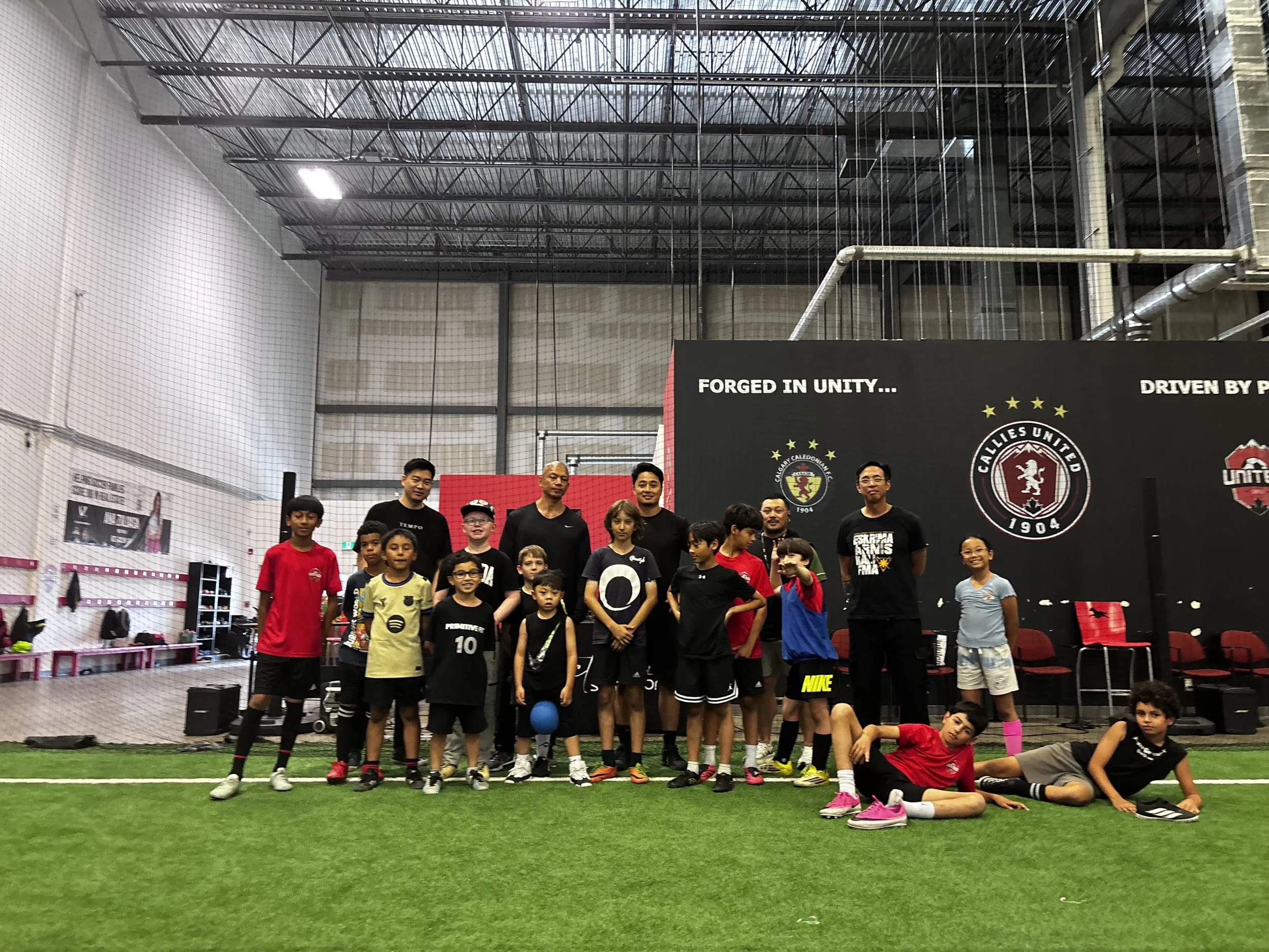 A group of children and coaches in sports attire posing for a photo inside an indoor sports facility with artificial turf and banners, including Calgary Callies United Soccer Club, in the background.