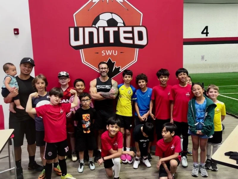 Group of kids and adults posing indoors in front of a large soccer-themed banner that says 'United SWU' with a soccer ball logo, some children wearing sports jerseys.