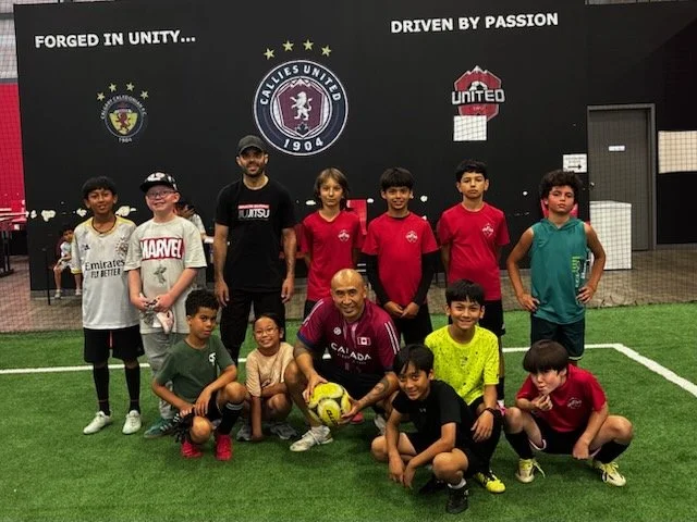 Group photo of young boys and a coach at a soccer field inside a stadium, with football gear, on a green turf, with banners and slogans on the wall.