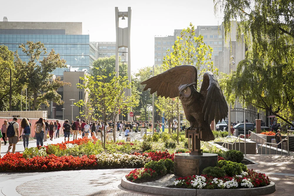 A city park with a bronze owl sculpture with wings spread, surrounded by colorful flowers and trees, with people walking in the background and modern office buildings nearby.