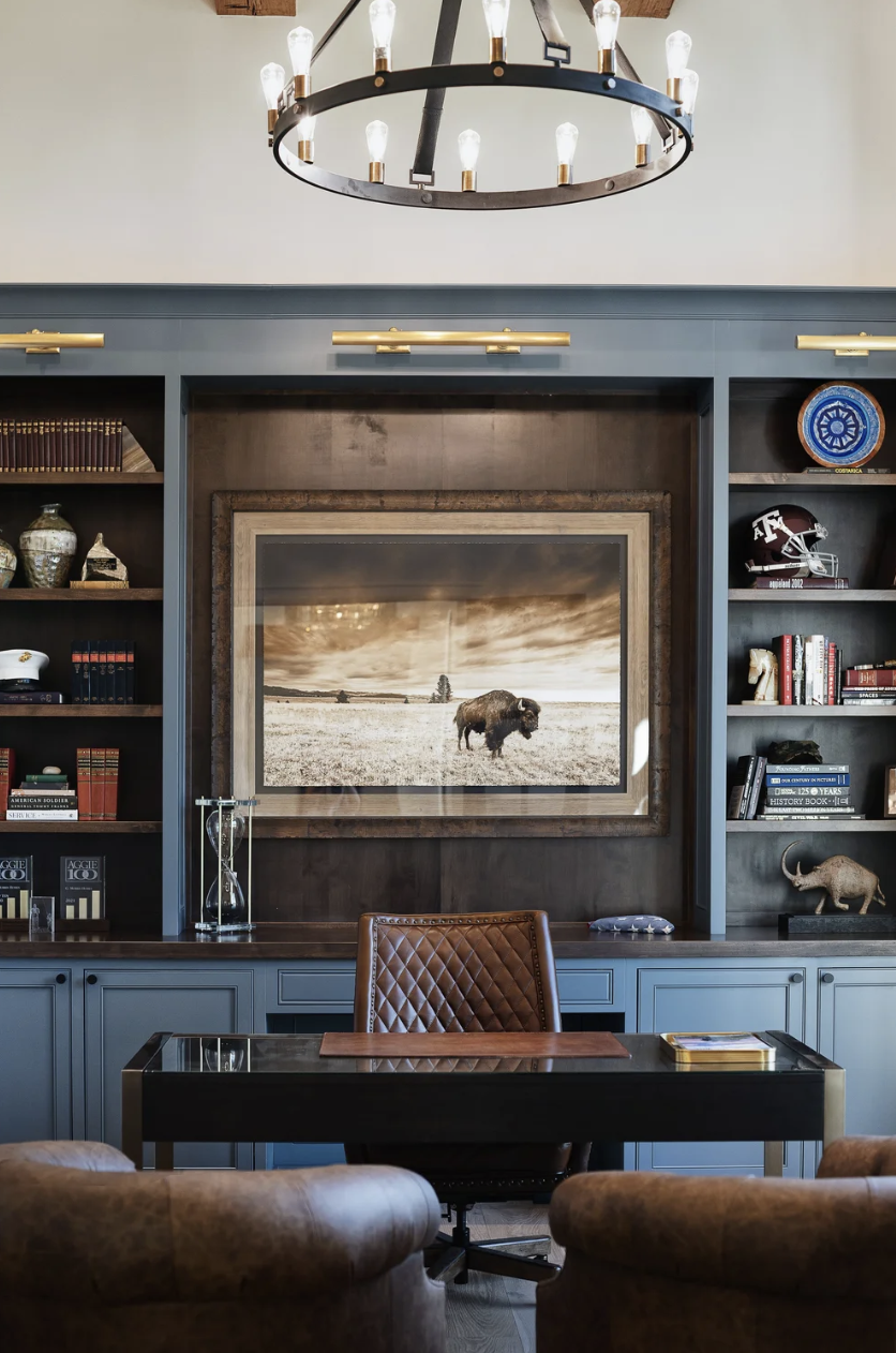 Moody home office with built-in shelving, blue cabinetry, leather seating, and framed buffalo artwork with brass lighting.