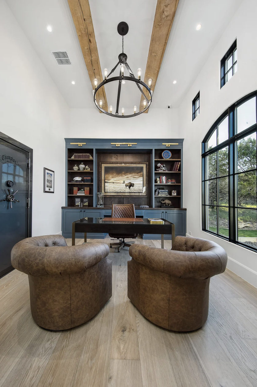 Refined home office featuring custom built-in shelving, blue cabinetry, leather seating, brass picture lighting, and framed buffalo artwork.