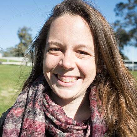 Close-up portrait of a smiling woman with long brown hair and a patterned scarf, standing outdoors in a sunny grassy area.