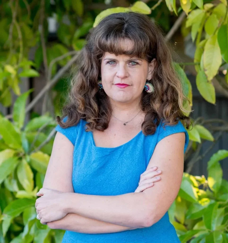 Portrait of Anat Shenker-Osorio with folded arms, wearing a blue dress, facing the camera.