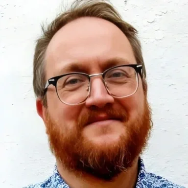 Headshot of a person with short light-brown hair, glasses and a red beard, wearing a patterned blue shirt against a white background