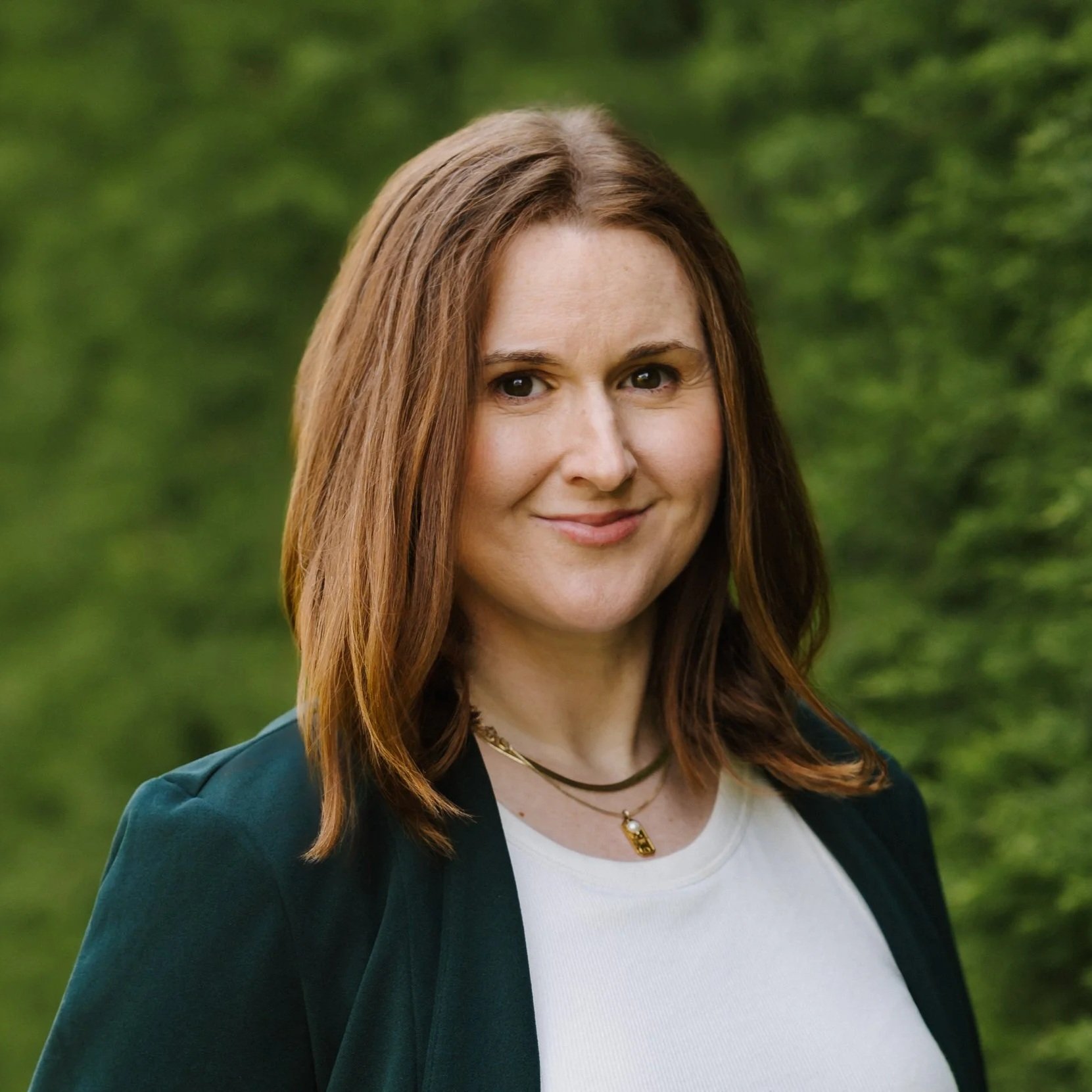 Woman with shoulder-length auburn hair smiling at the camera, wearing a dark green blazer and white top, standing outdoors beside a lush green hedge.