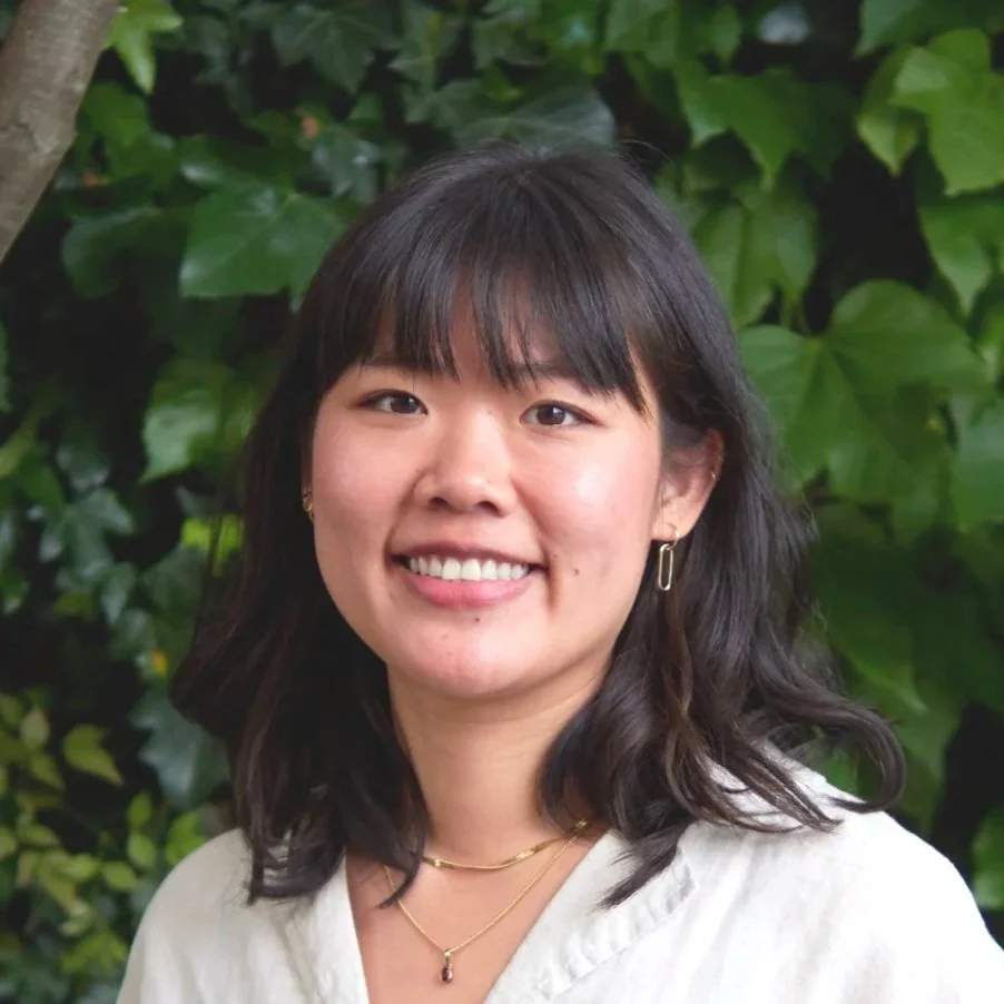 A smiling woman in a white linen shirt stands outdoors in front of lush green foliage and trees.
