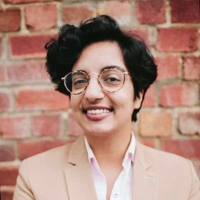 A smiling person with short dark hair and round glasses stands in front of a red brick wall, wearing a light-colored blazer over a pale shirt. They are facing the camera with a relaxed, friendly expression.
