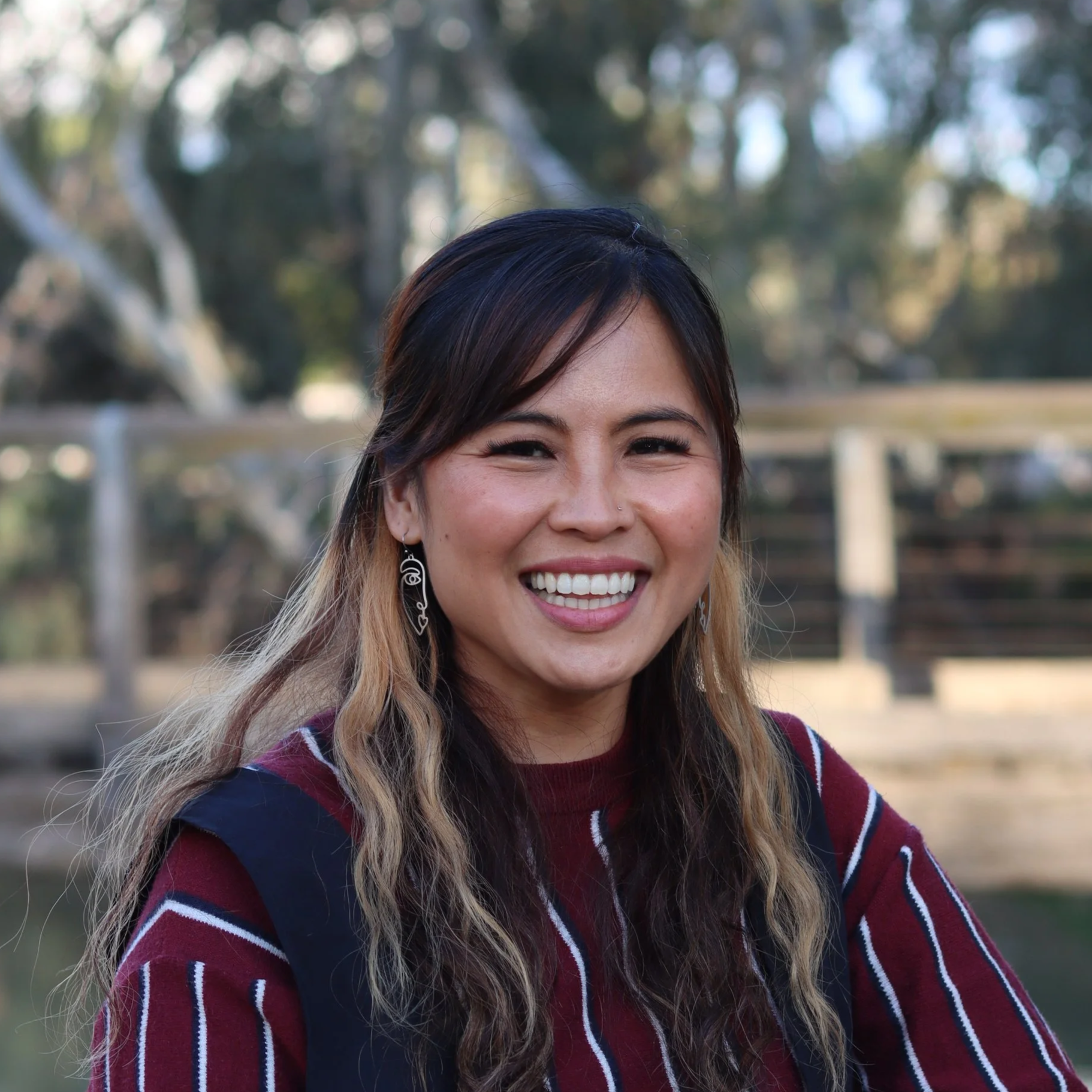 A smiling woman stands outdoors by a wooden railing beside a river, wearing a burgundy striped top and dark overalls, with trees and water in the background.