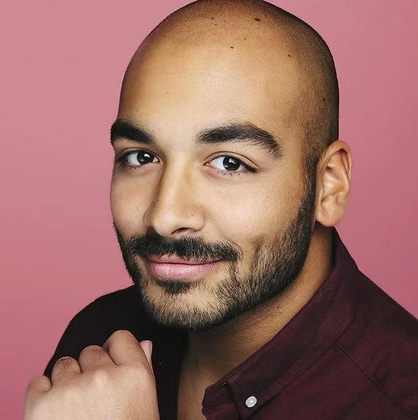 Close-up headshot of a smiling person with a shaved head and neatly trimmed beard, resting their chin on their hand against a pink background.