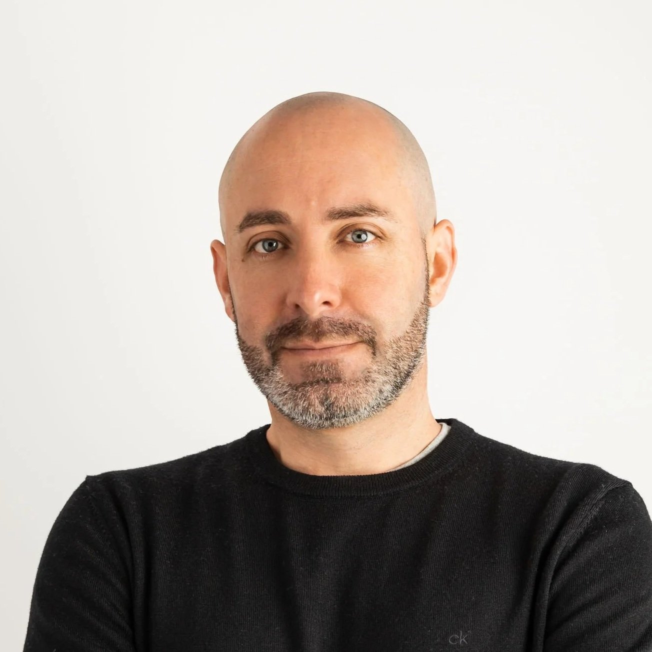 Bald man with a short beard looking at the camera with his arms crossed, wearing a black long-sleeve top against a plain white background.