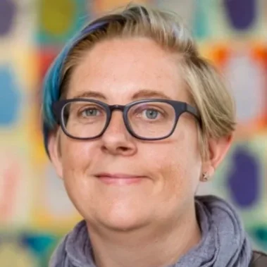 Headshot of a person with short blonde hair and glasses, wearing a grey scarf, with a colourful blurred background.