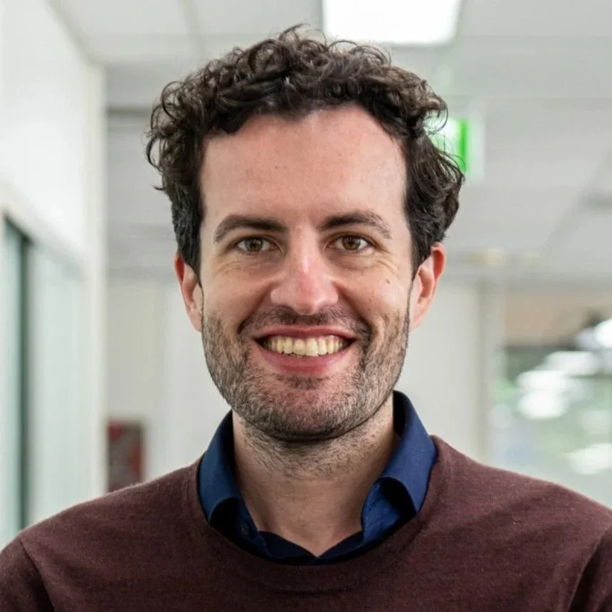 A smiling man with short curly dark hair and light stubble, wearing a dark shirt and brown sweater, standing indoors in a softly lit office hallway.