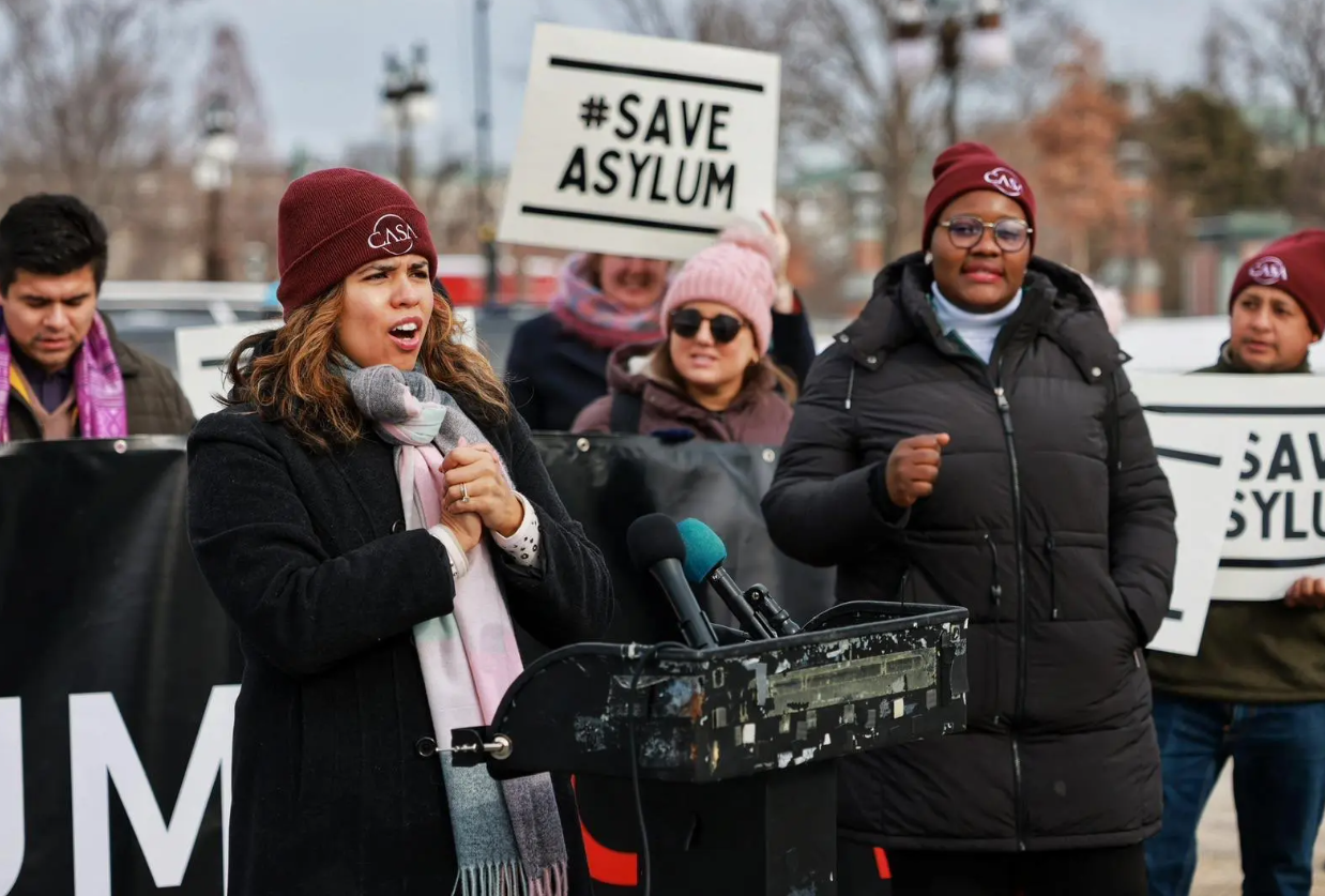 Photo of Jossie Sapunar speaking. There is a poster held in the background that says '#SAVE ASYLUM'