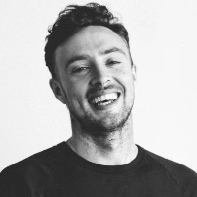 Black-and-white portrait of a smiling man with short dark hair and light facial hair, wearing a dark crew-neck top against a plain white background.