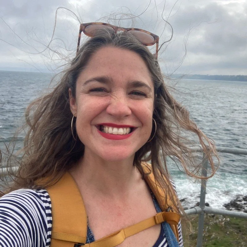 Picture of Natasha in front of the sea with a big smile & messy hair blowing in the wind
