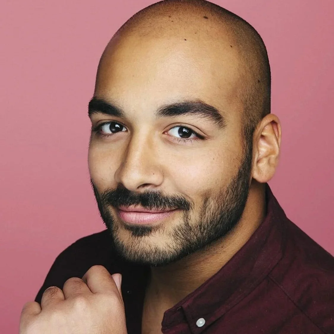 Close-up portrait of a smiling man with a shaved head and short dark beard, wearing a burgundy shirt against a pink background.