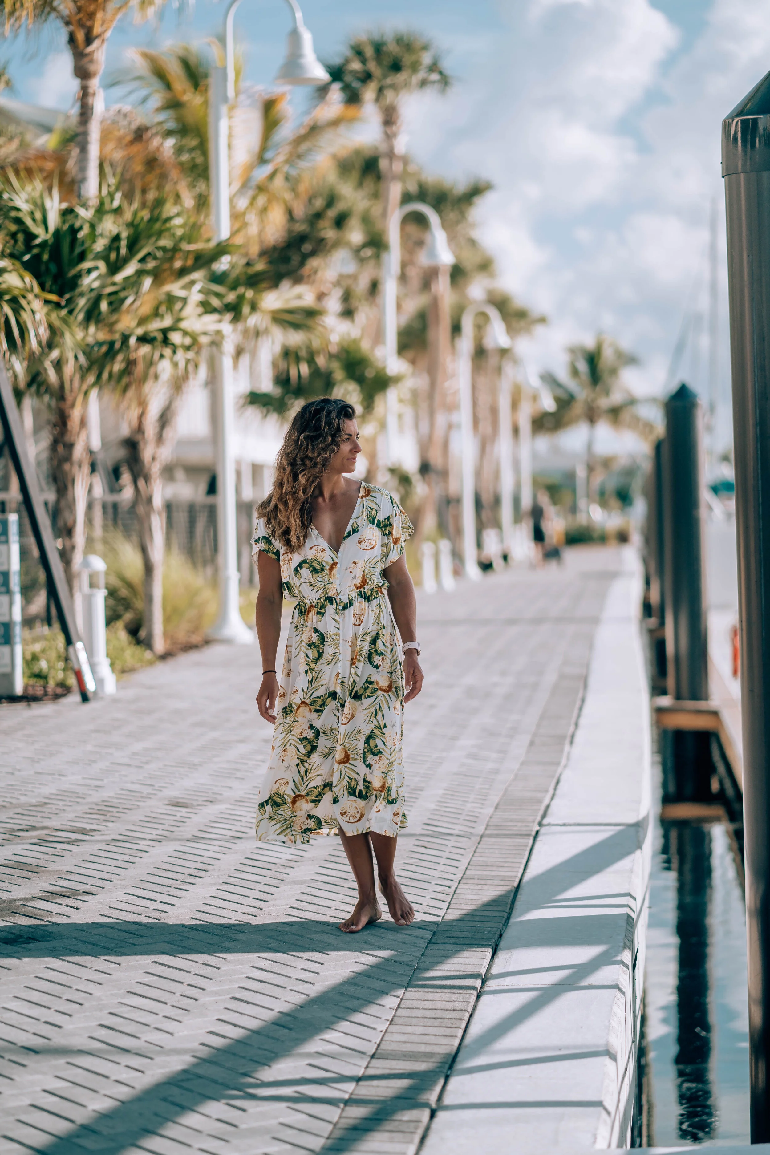 A woman walking barefoot on a marina dock, wearing a floral dress with palm trees and boats in the background on a sunny day.