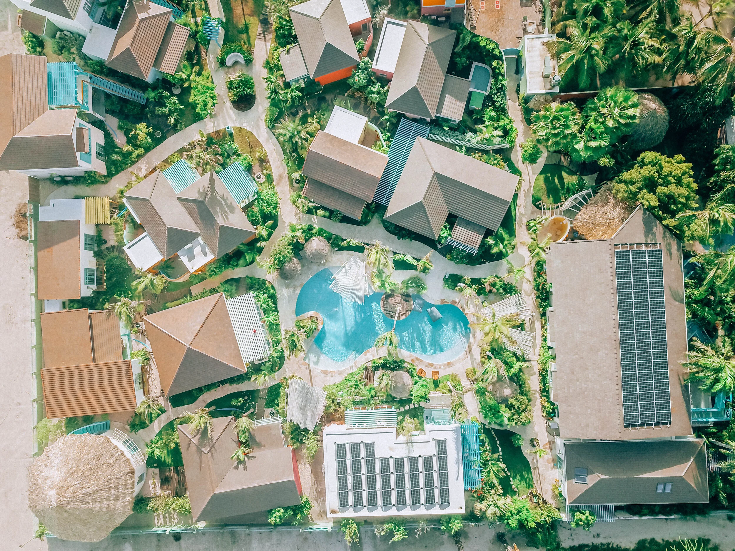 Aerial view of a tropical resort with multiple cottages, a swimming pool, lush greenery, and palm trees.