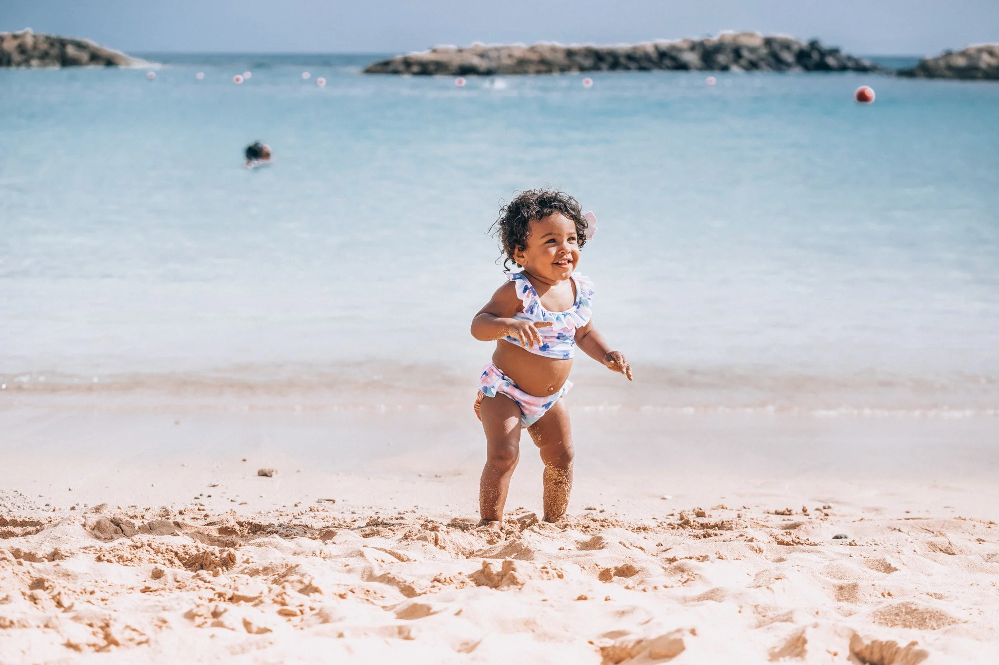 A young girl running on a sandy beach near the ocean, wearing a ruffled swimsuit.