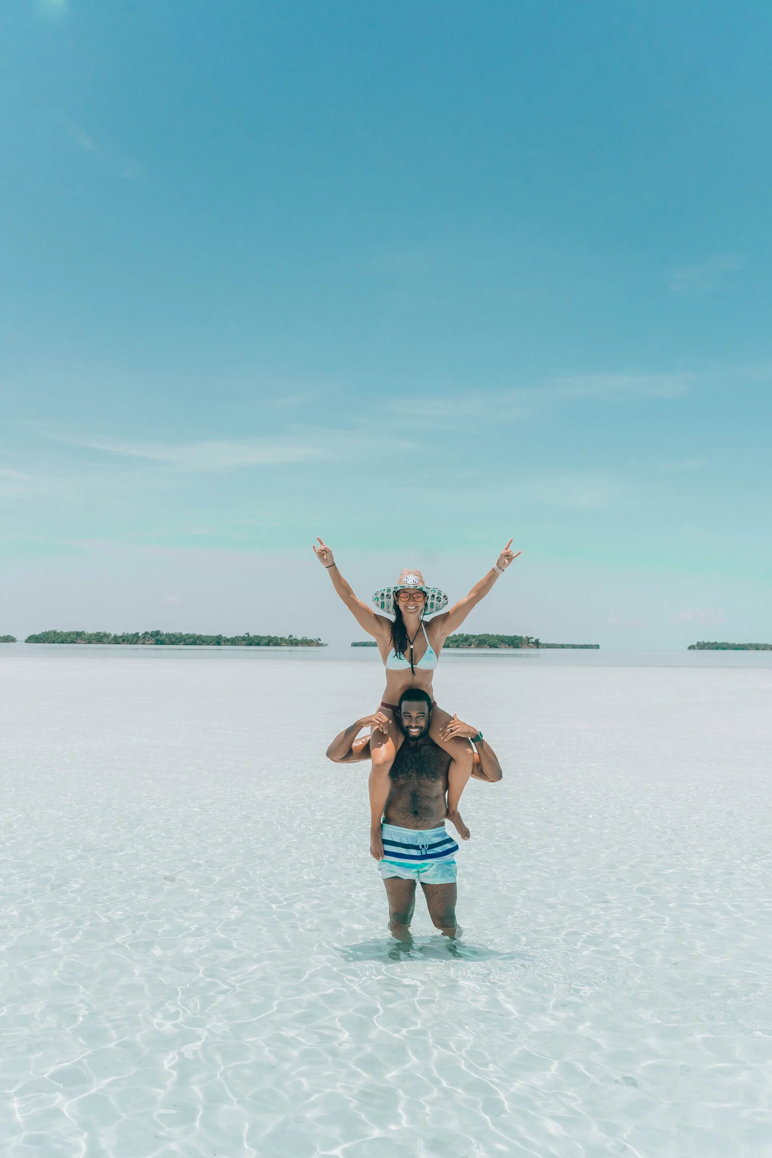 A man and woman enjoying a day at the beach, with the woman sitting on the man's shoulders, both making peace signs. The water is clear and shallow, and there are islands in the background under a blue sky.
