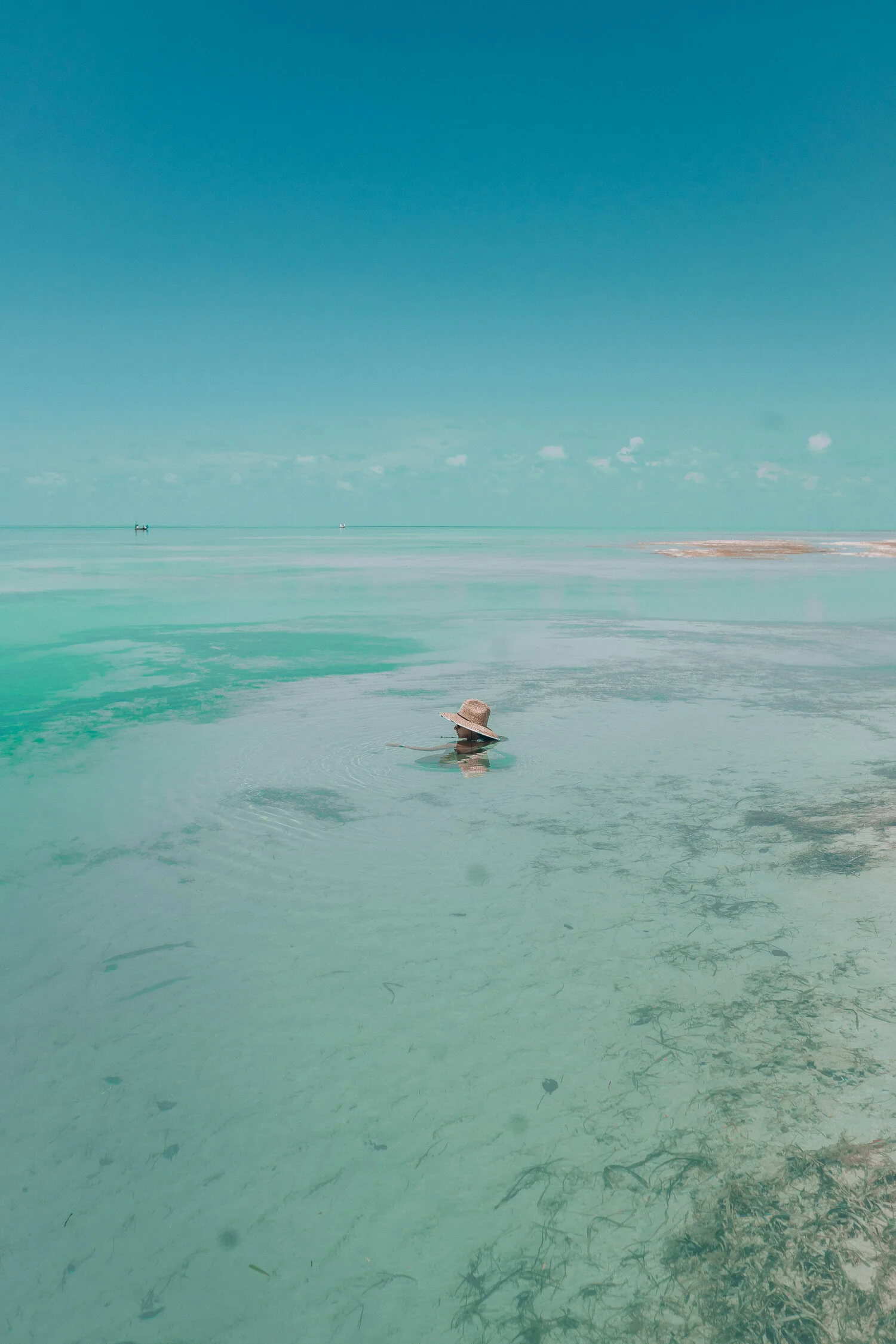 A person in a large straw hat swimming in a shallow, calm, turquoise body of water with a clear blue sky above and distant boats in the background.
