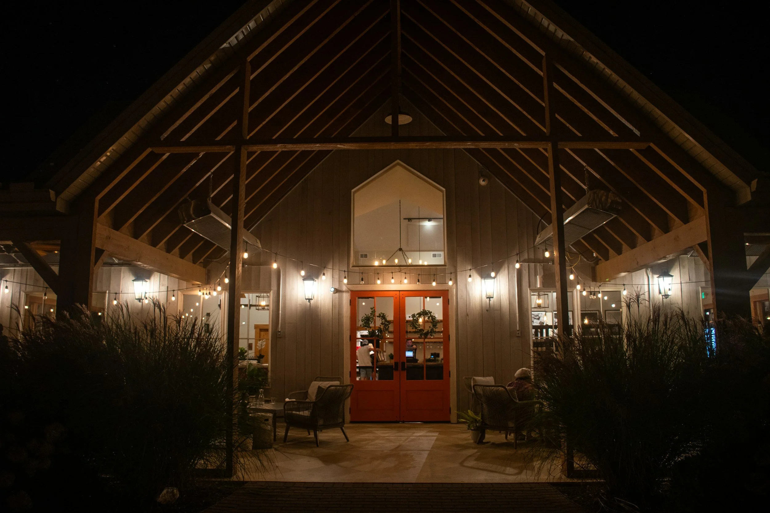 Night view of a warmly lit outdoor patio and entrance of a restaurant or cafe, with patio chairs, hanging string lights, and wooden architecture.