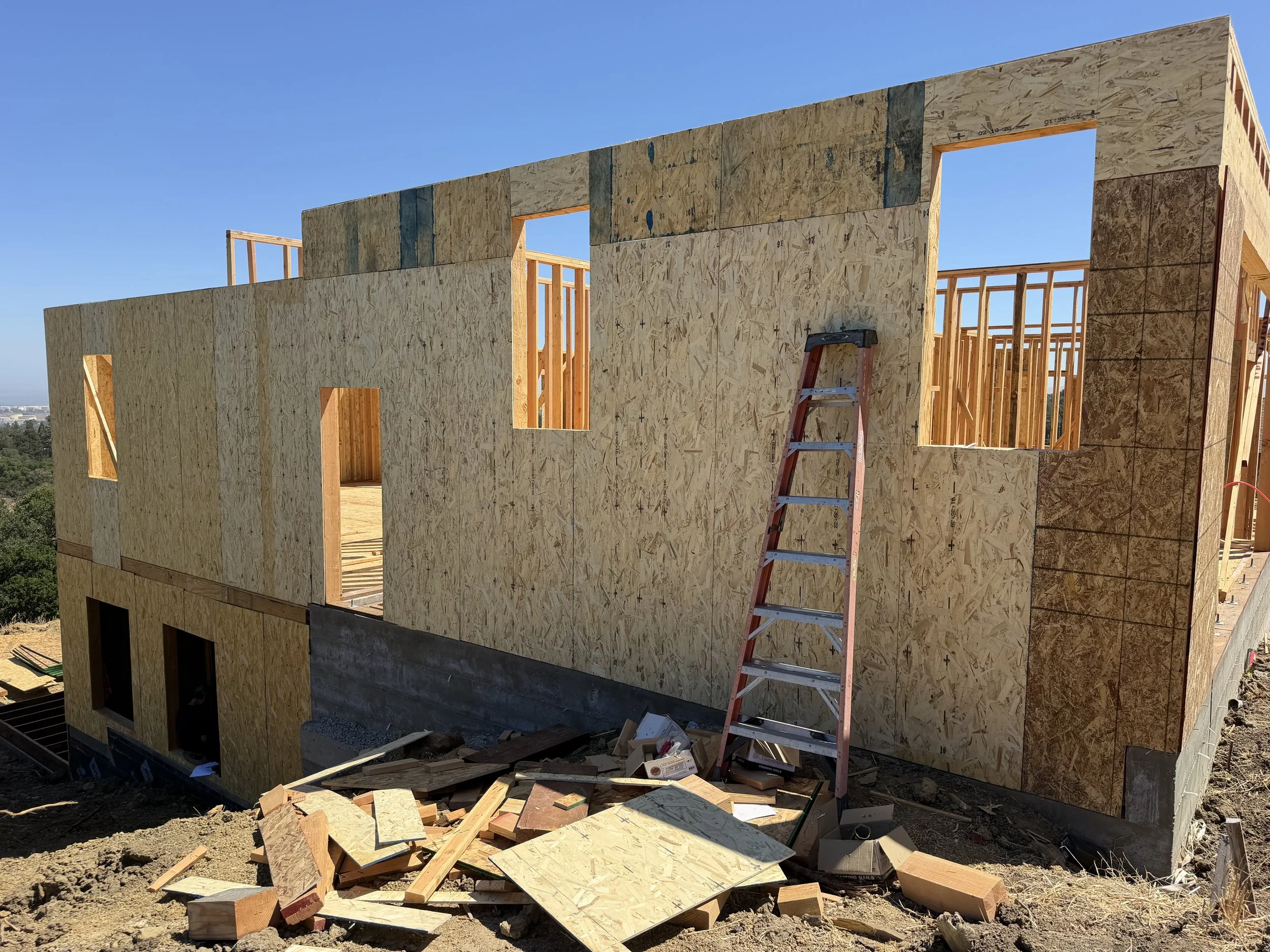 The image shows the wooden frame of a building under construction, with exterior walls partially covered with oriented strand board (OSB) sheathing. There is a red ladder leaning against the wall, and construction tools and debris are scattered on the ground below. The sky is clear and blue.