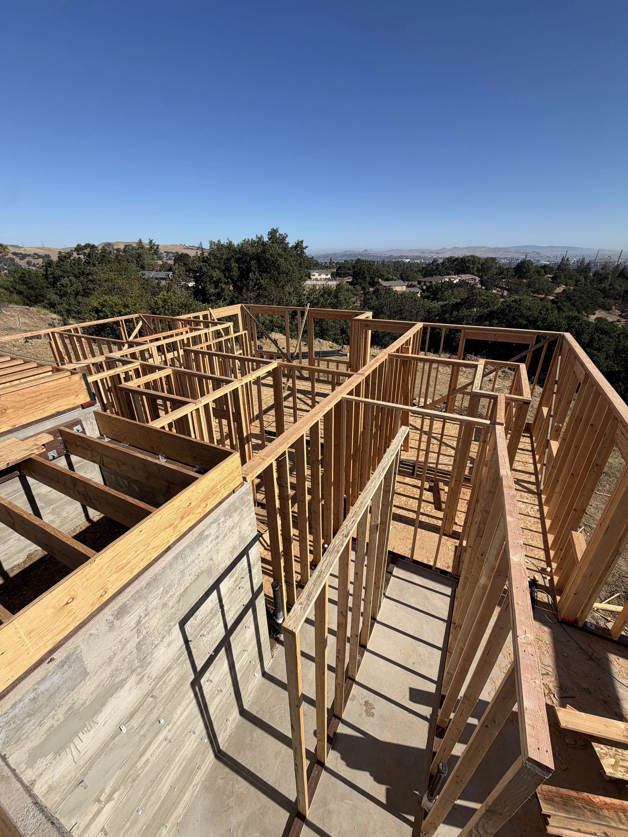 Wooden framing for a building under construction on a hillside with a view of trees, houses, and distant mountains under a clear blue sky.