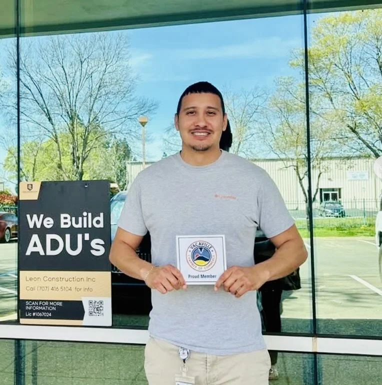 A man standing inside a building holding a 'Proud Member' badge. Behind him, print ads display 'We Build ADU's' and construction company details, with large glass windows showing trees and a parking lot outside.