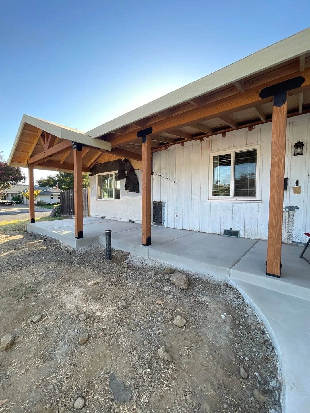 A house with a new concrete porch and wooden posts supporting a roof extension, surrounded by dirt and rocks.