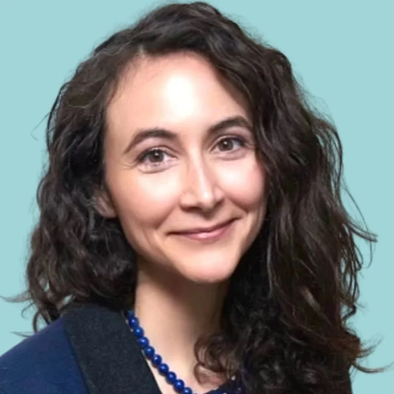 A woman with shoulder-length curly brown hair and light skin smiling, wearing a dark top and a blue beaded necklace.