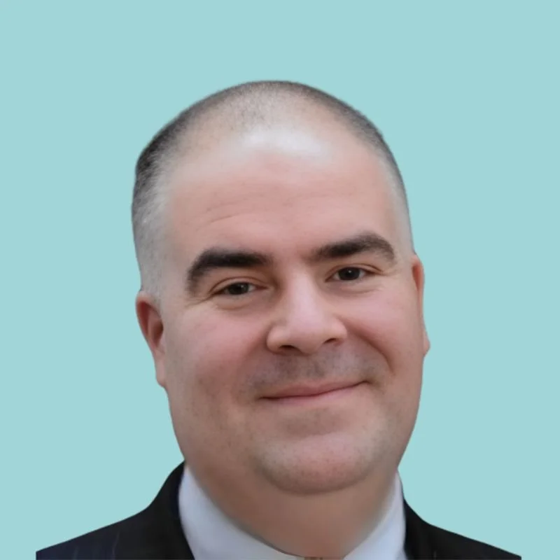 Close-up of a man with short hair, dressed in a dark suit, white shirt, and tie, smiling against a gray background.