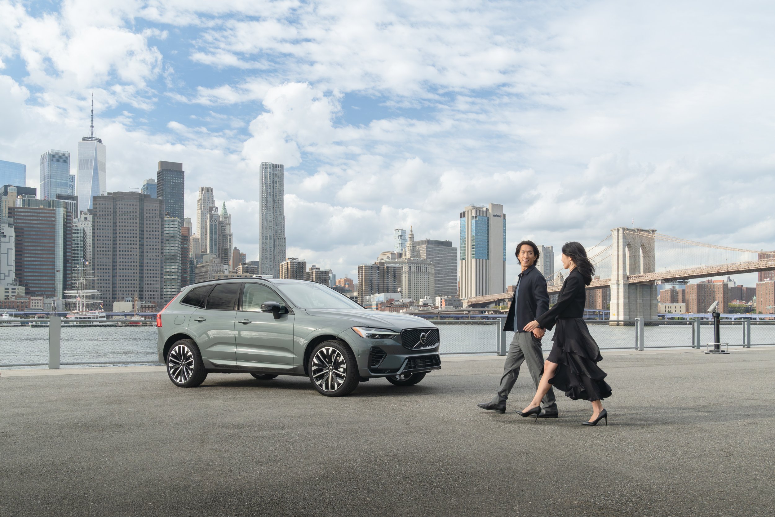 A man and woman walking hand-in-hand along a waterfront with a city skyline and a bridge in the background. There is a gray SUV parked nearby.
