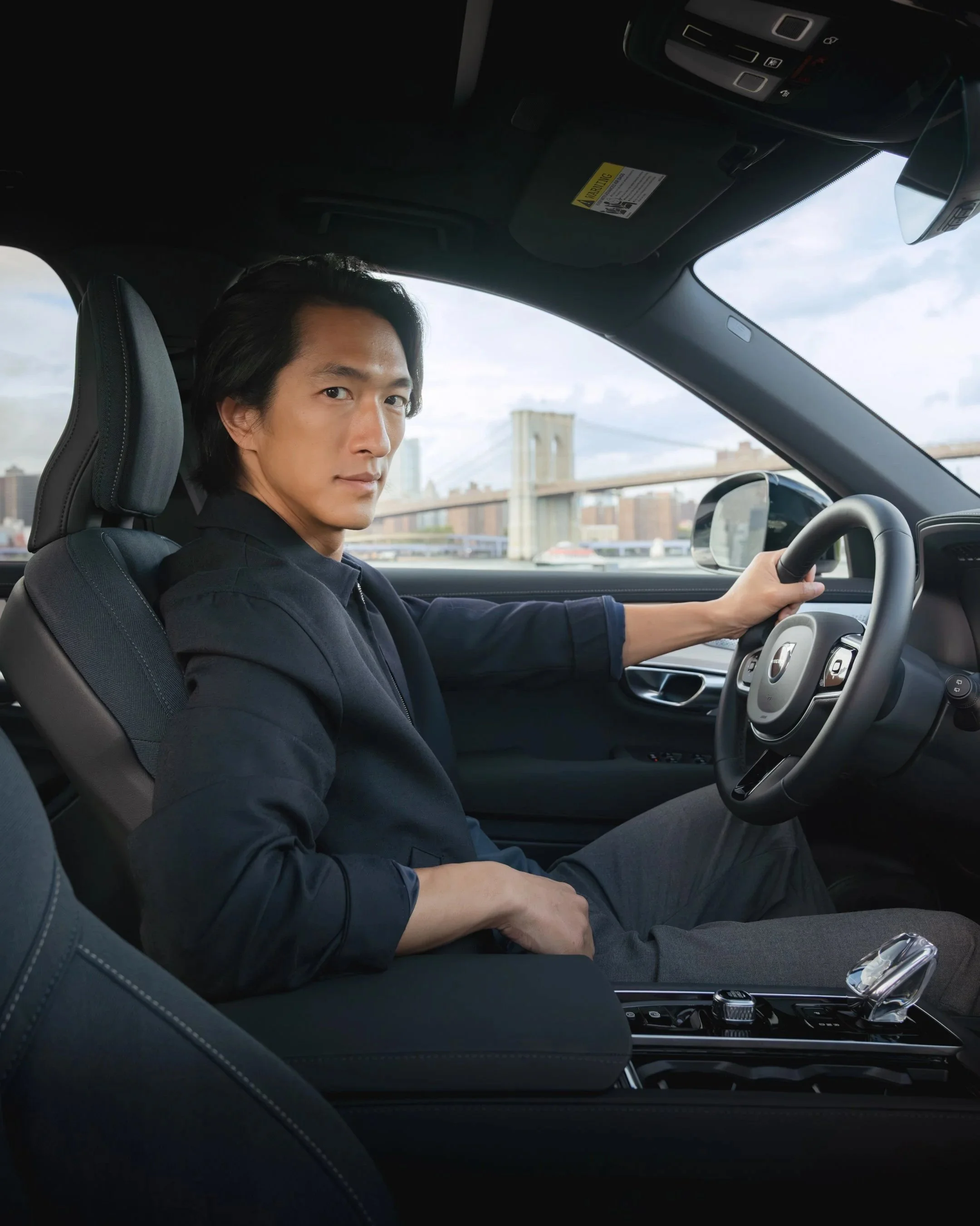 A man sitting in a car, holding the steering wheel, with a cityscape and bridge visible through the window.