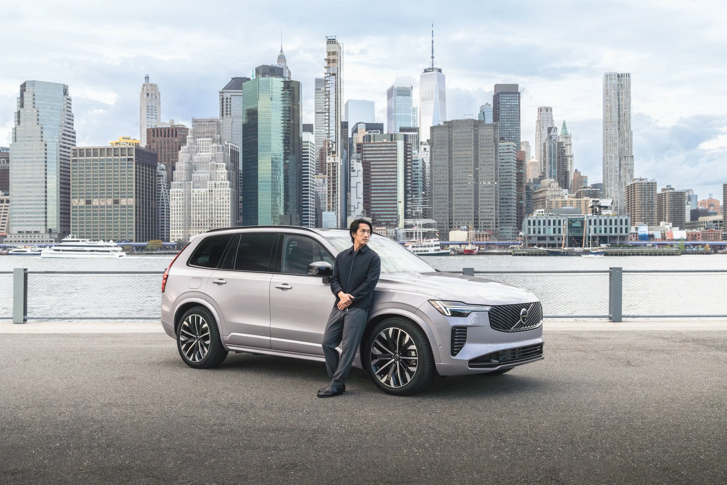 A man leaning against a silver Volvo SUV with a city skyline background over water, cloudy sky.