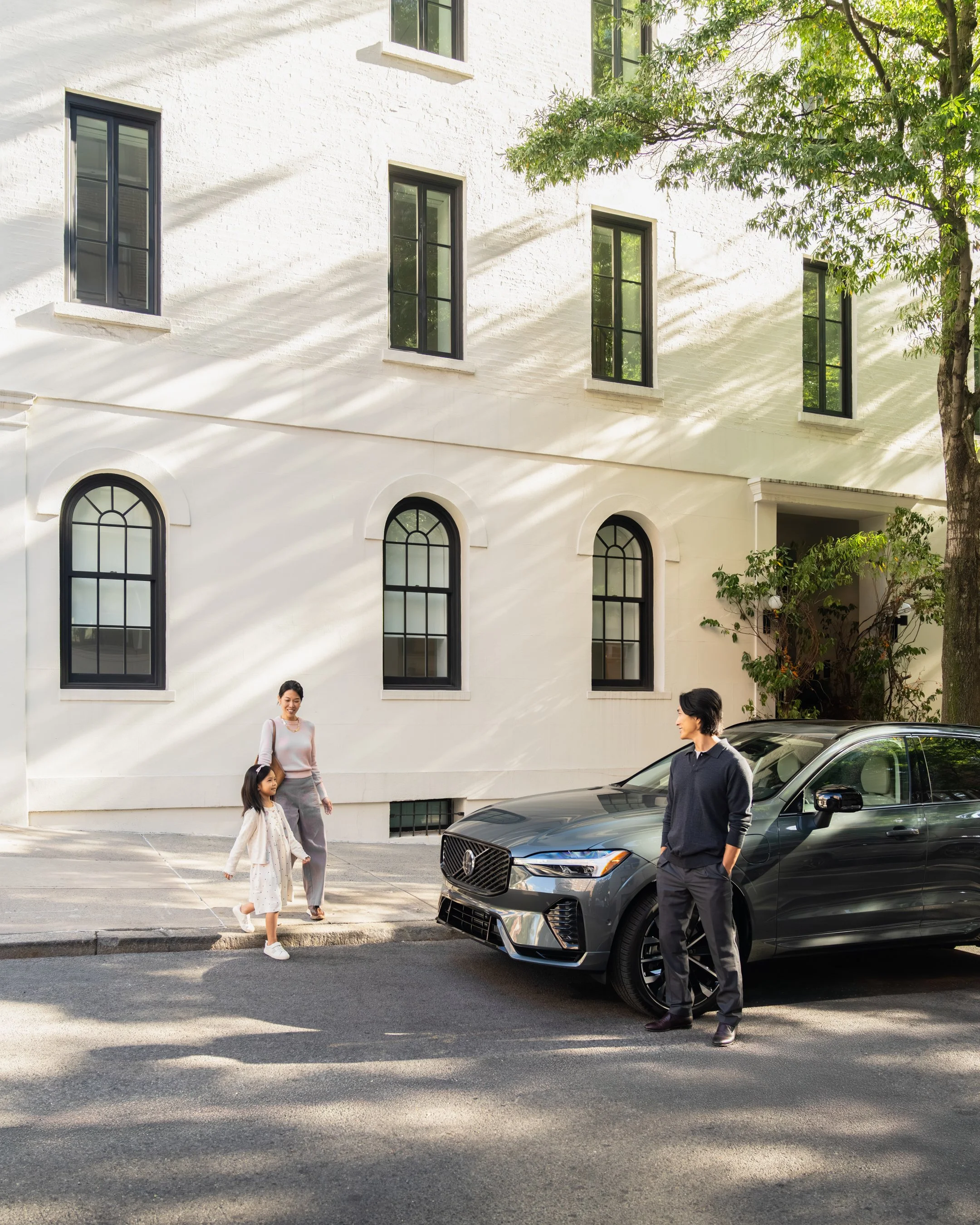 A family with a woman, young girl, and man near a dark gray car parked on a city street in front of a white building with large windows and trees, on a sunny day.