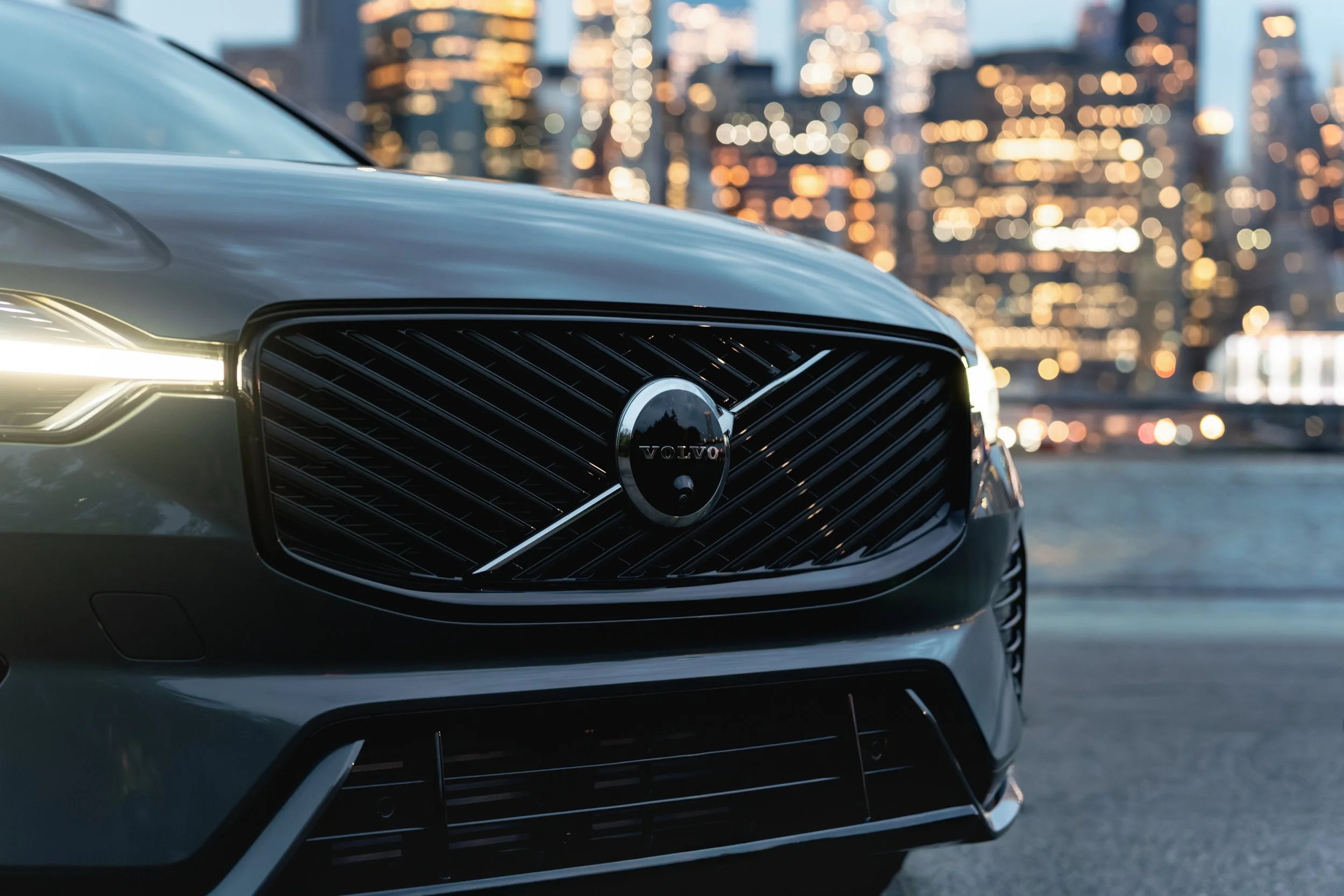 Close-up of a Volvo car's front grille with city skyscrapers in the blurred background during dusk.