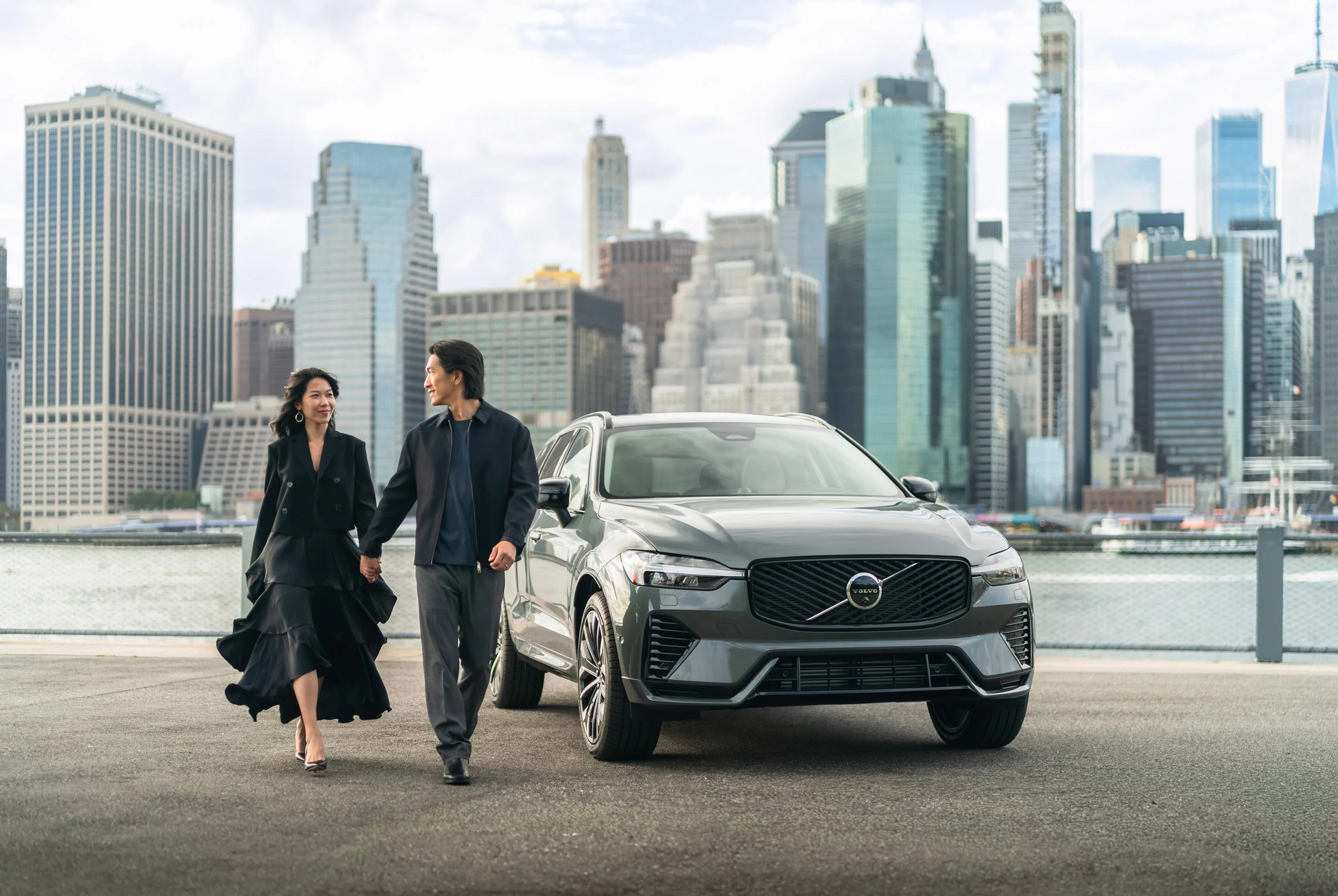A man and woman walking hand in hand with a gray Volvo car parked nearby in an urban setting with high-rise buildings in the background