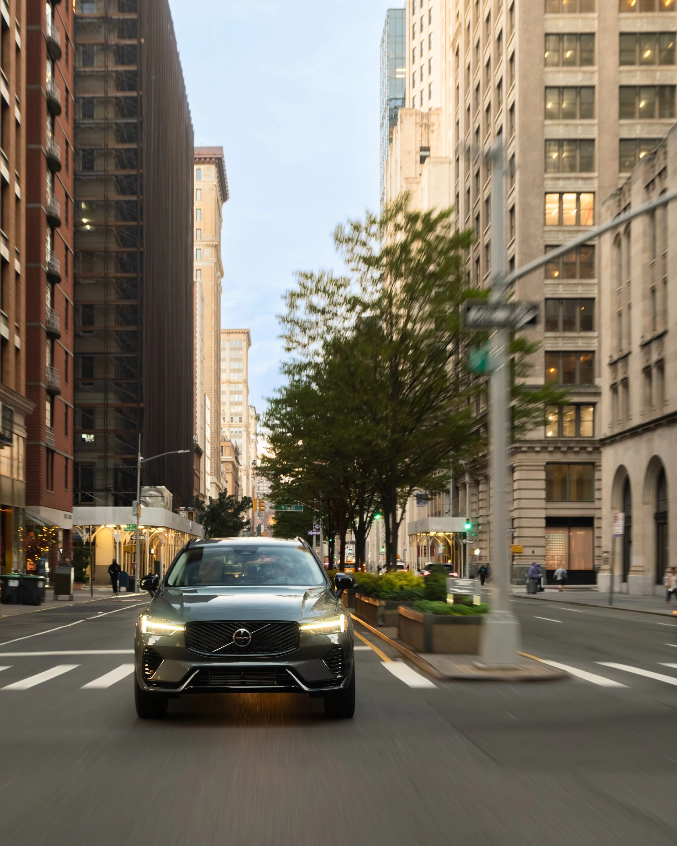 A black Volvo SUV driving down a city street surrounded by tall buildings, with trees and pedestrians nearby.