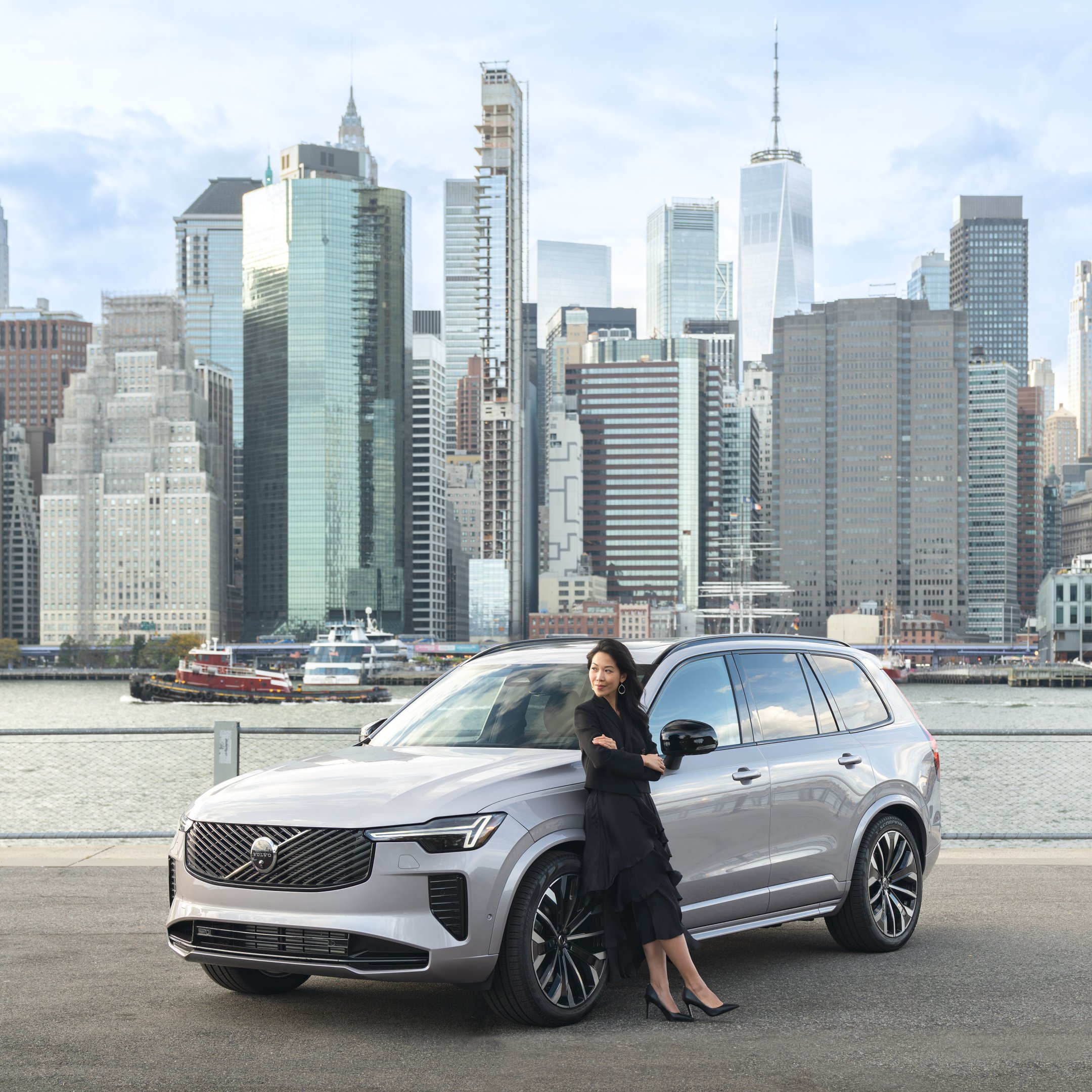 A woman standing beside a silver SUV on a city street with a skyline of skyscrapers in the background.