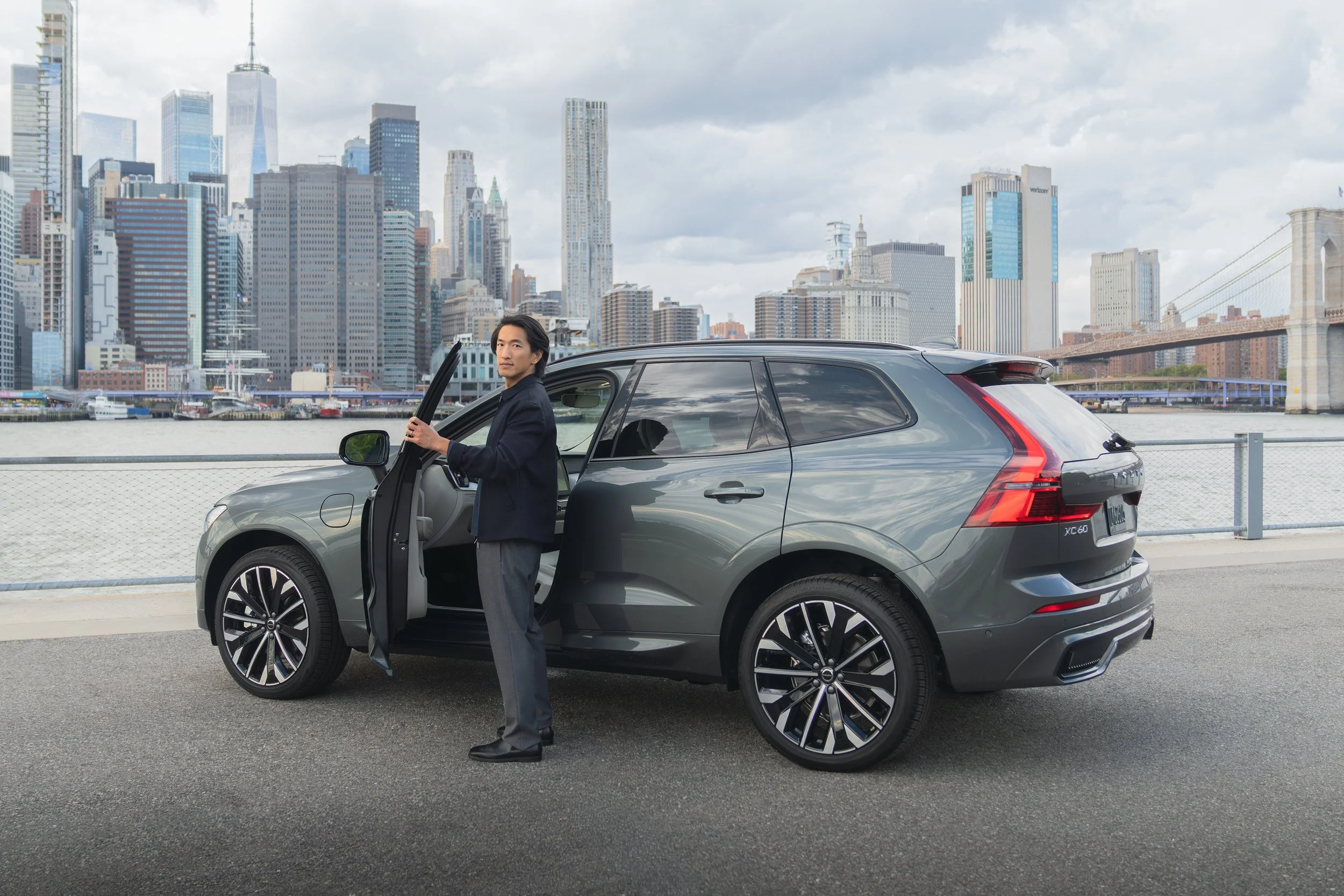A man in a dark jacket and gray trousers standing next to a gray SUV by the water, with a city skyline of skyscrapers and a bridge in the background.