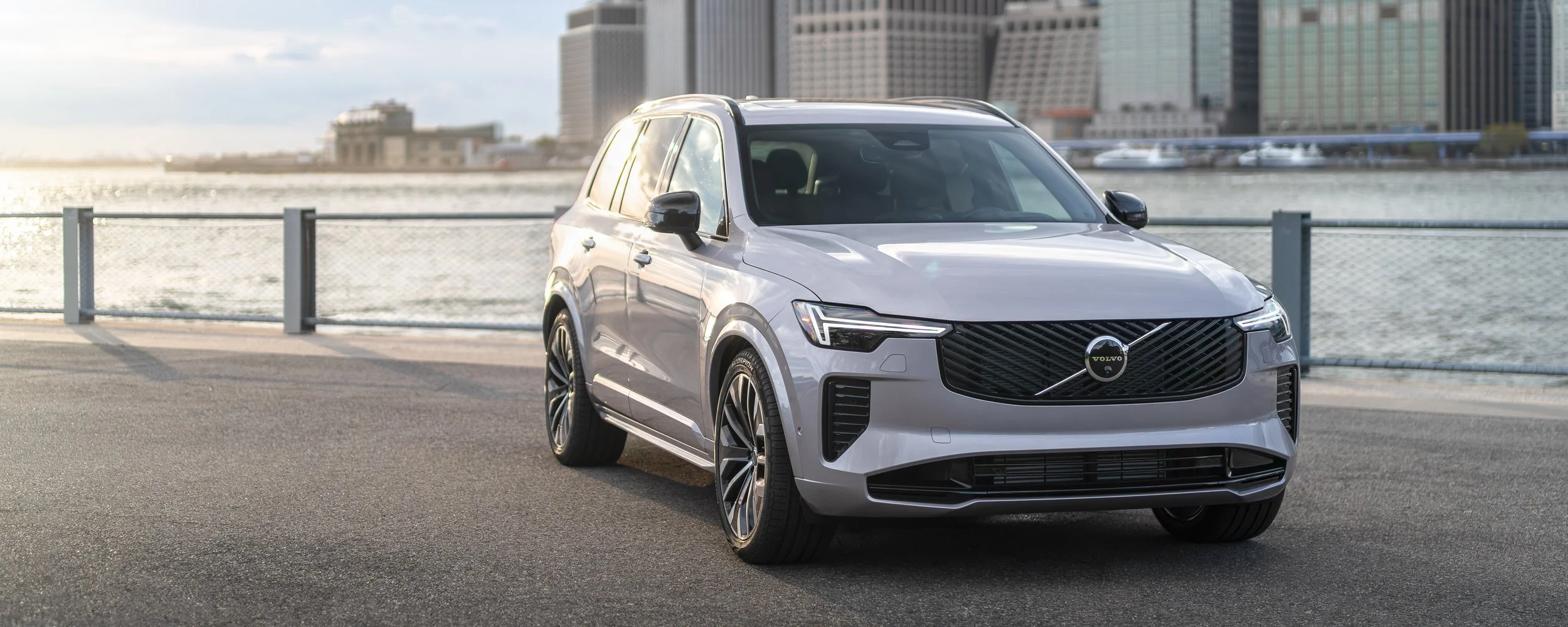 Silver Volvo SUV parked near a waterfront with city buildings in the background during sunset.