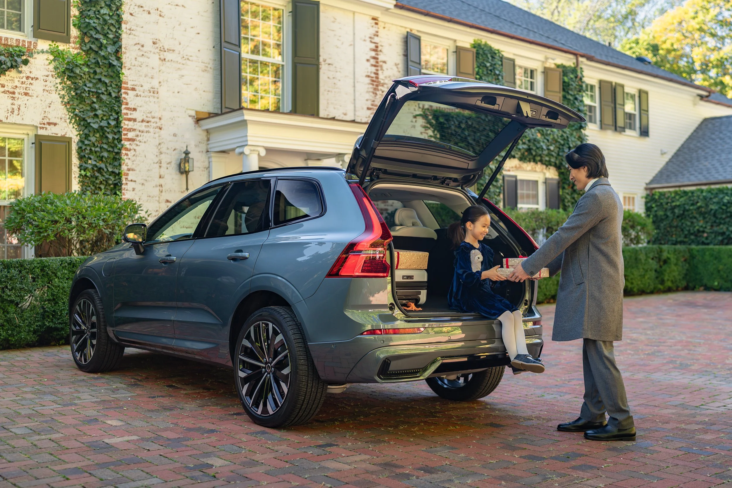 A woman is handing a gift box to a girl sitting in the trunk of a blue SUV, with houses and greenery in the background.