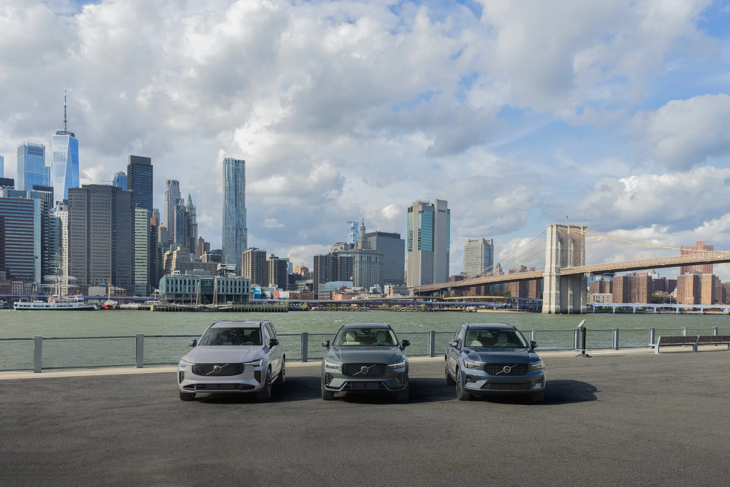 Three gray cars parked side by side on a paved area overlooking the water and city skyline, with a bridge in the background under a partly cloudy sky.