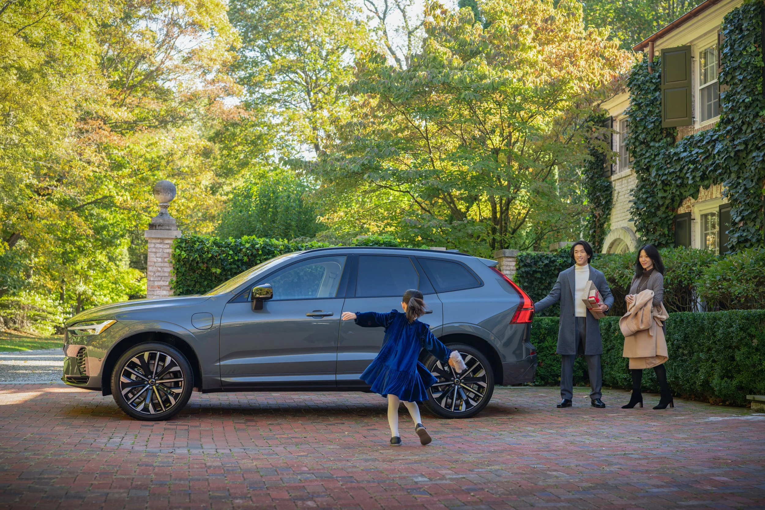 A family is standing near a parked dark gray SUV on a brick driveway. A young girl in a blue dress is running towards the car to open the door. A man and woman are smiling and holding shopping bags, ready to get into the vehicle, with a background of