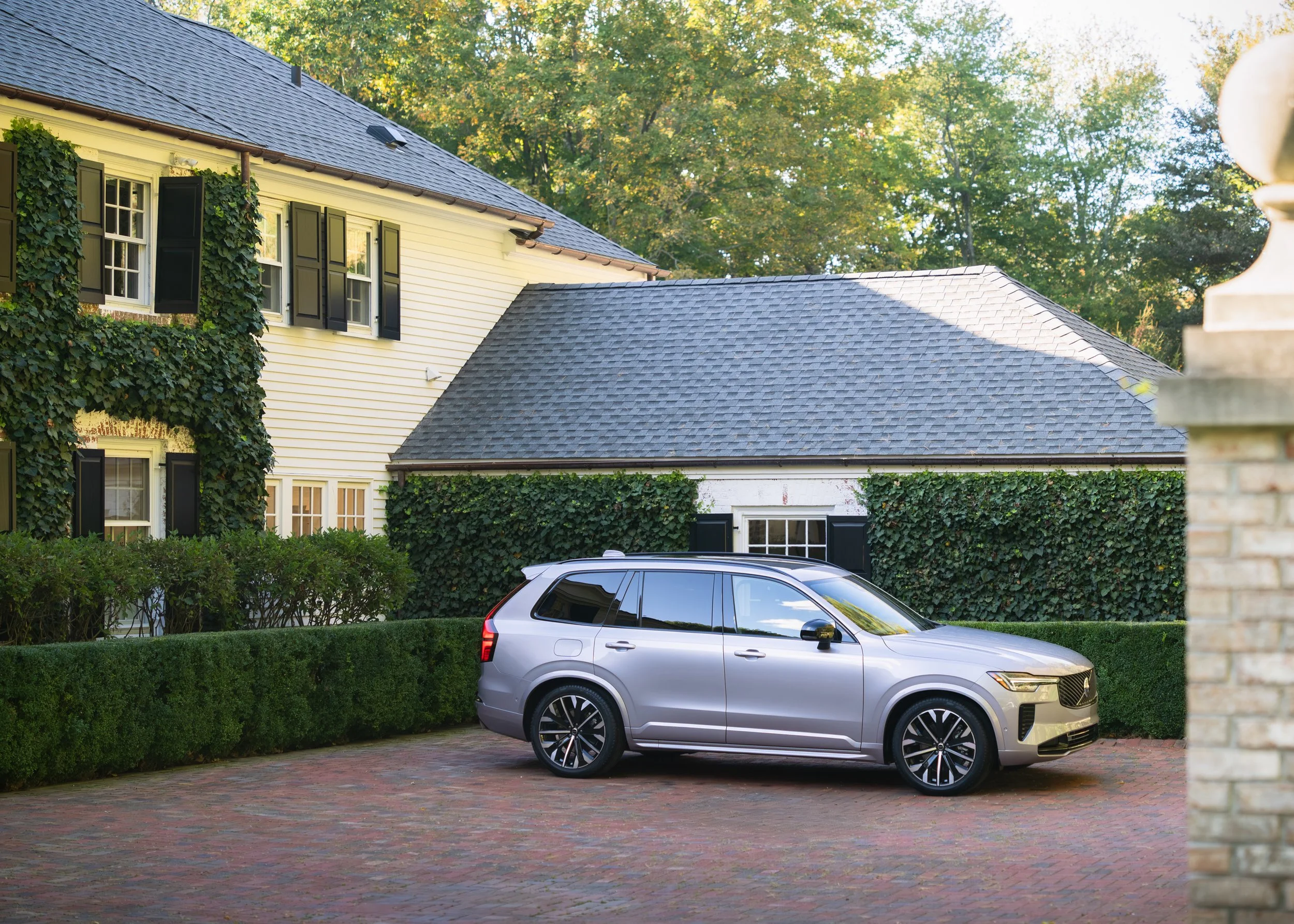 A silver SUV parked on a brick driveway in front of a house with beige siding, black window shutters, and ivy-covered walls, surrounded by green trees.