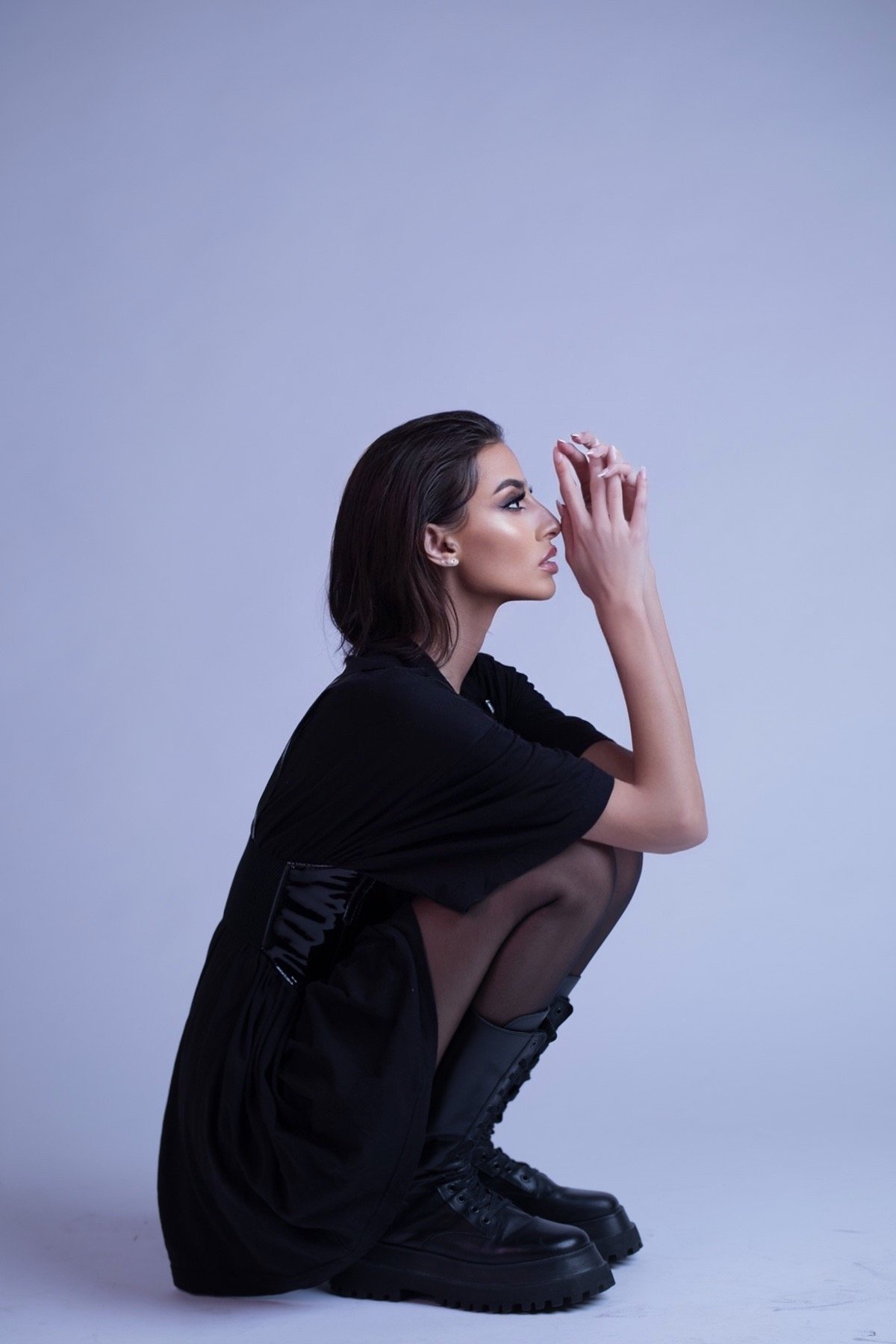 A person squatting sideways in a thoughtful pose, wearing a black dress and black combat boots against a light blue background.