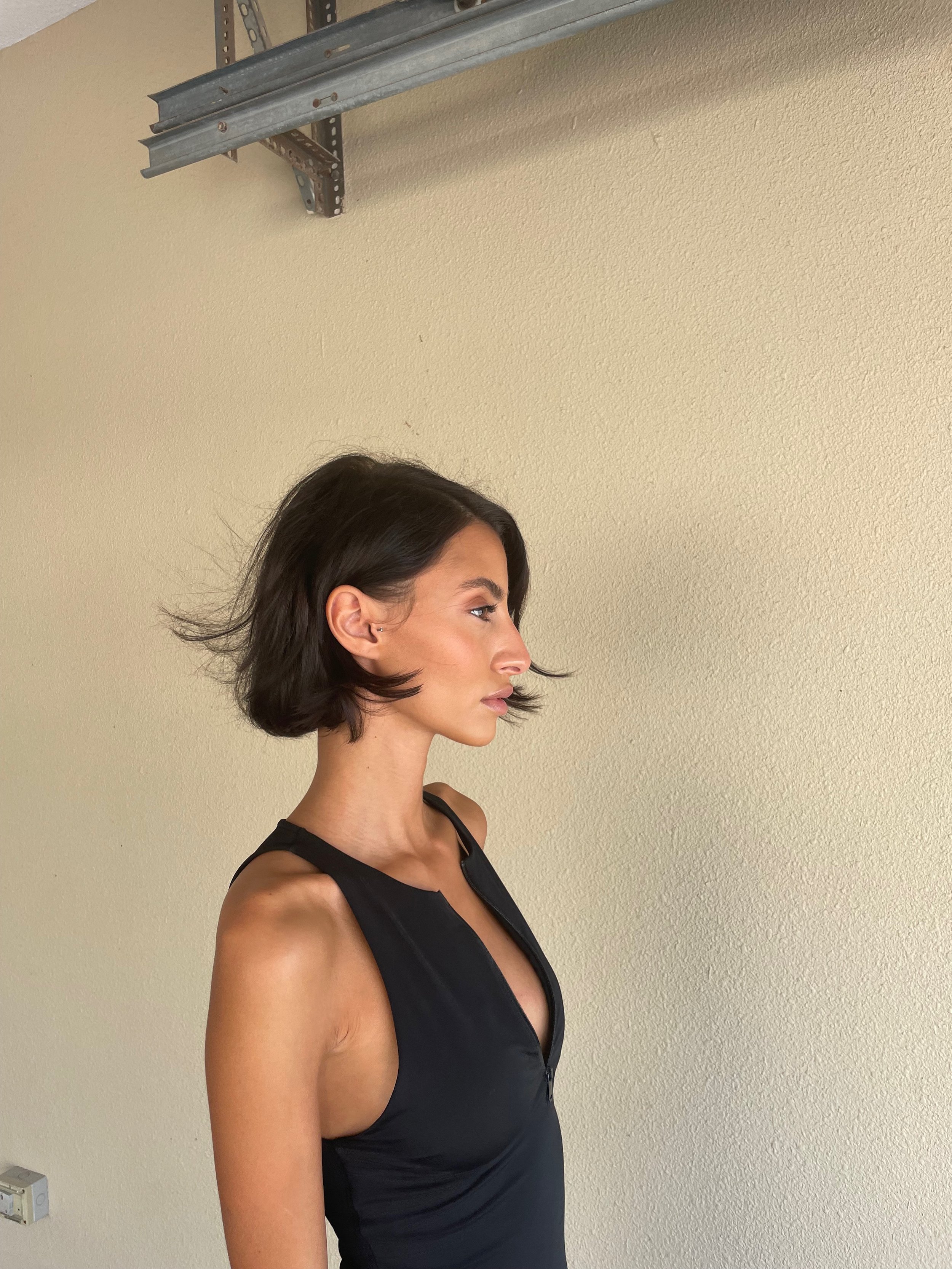 Woman with short hair in a black dress standing against a beige wall, with piping visible above.
