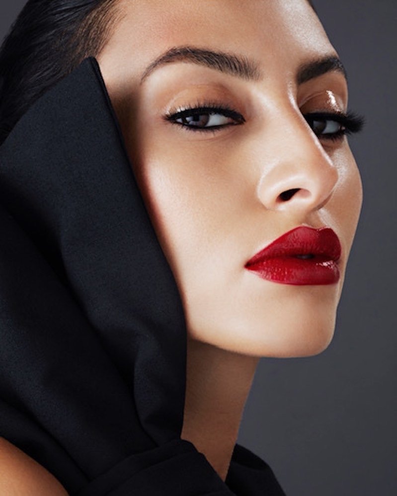 Close-up of a person with bold red lipstick, wearing a black hooded garment, neutral makeup, and styled black hair.